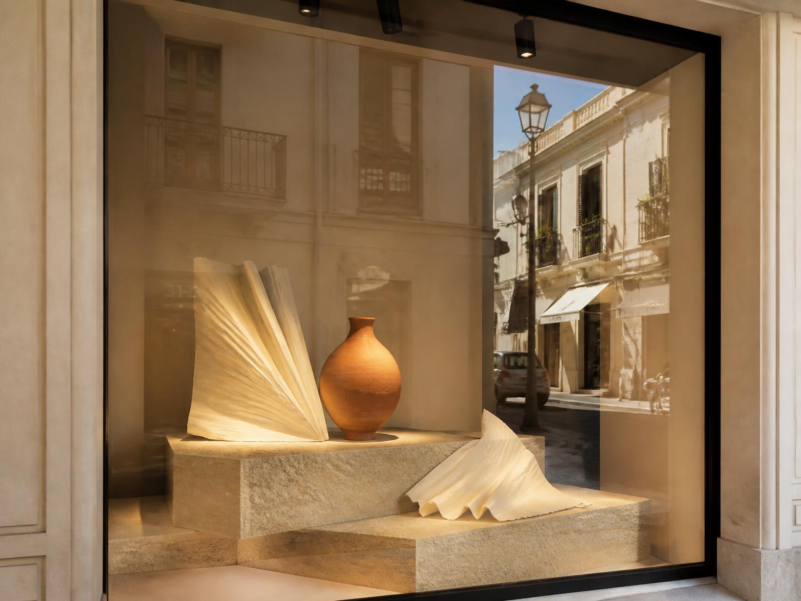 Escaparate minimalista con peanas de piedra, formas de papel plegado y vasija de terracotta